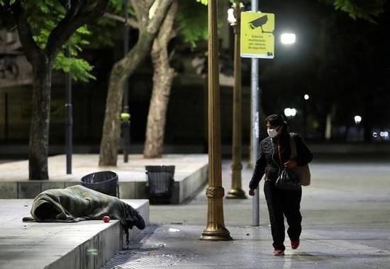 (图说:2020年4月,阿根廷首都布宜诺斯艾利斯街头,一位戴着口罩的妇女从一位无家可归者身边走过。图/Reuters)